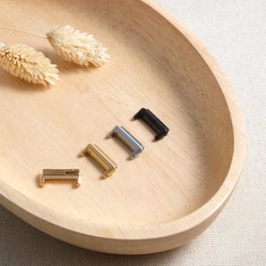 May include: A collection of metal watch band clasps in gold, silver, and black, arranged on a light wooden tray. The clasps are rectangular with a curved design. Two dried wheat stalks are also on the tray.