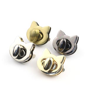 Könnte beinhalten: Vier metallene katzenförmige Verschlüsse in Gold-, Silber- und Gunmetal-Ausführungen. Jeder Verschluss hat ein Katzengesicht-Design mit einem zentralen, oval geformten Verschluss. Die Verschlüsse sind für Taschen oder andere Bastelprojekte konzipiert.