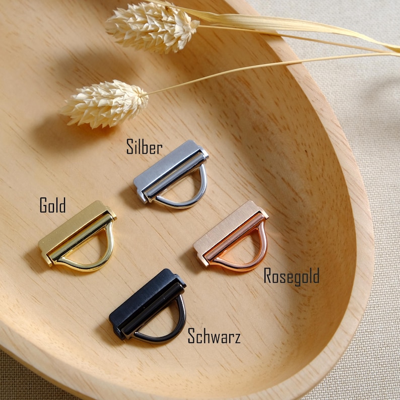 May include: Four metal purse hardware pieces in gold, silver, rose gold, and black, each with a rectangular bar and a curved handle. The pieces are arranged on a wooden tray with dried floral accents. The text on the image indicates the colour of each piece.