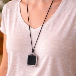 May include: A black smartwatch with a silver case hangs from a black cord necklace. The watch has a square face and is worn around the neck. The person is wearing a white v-neck top.