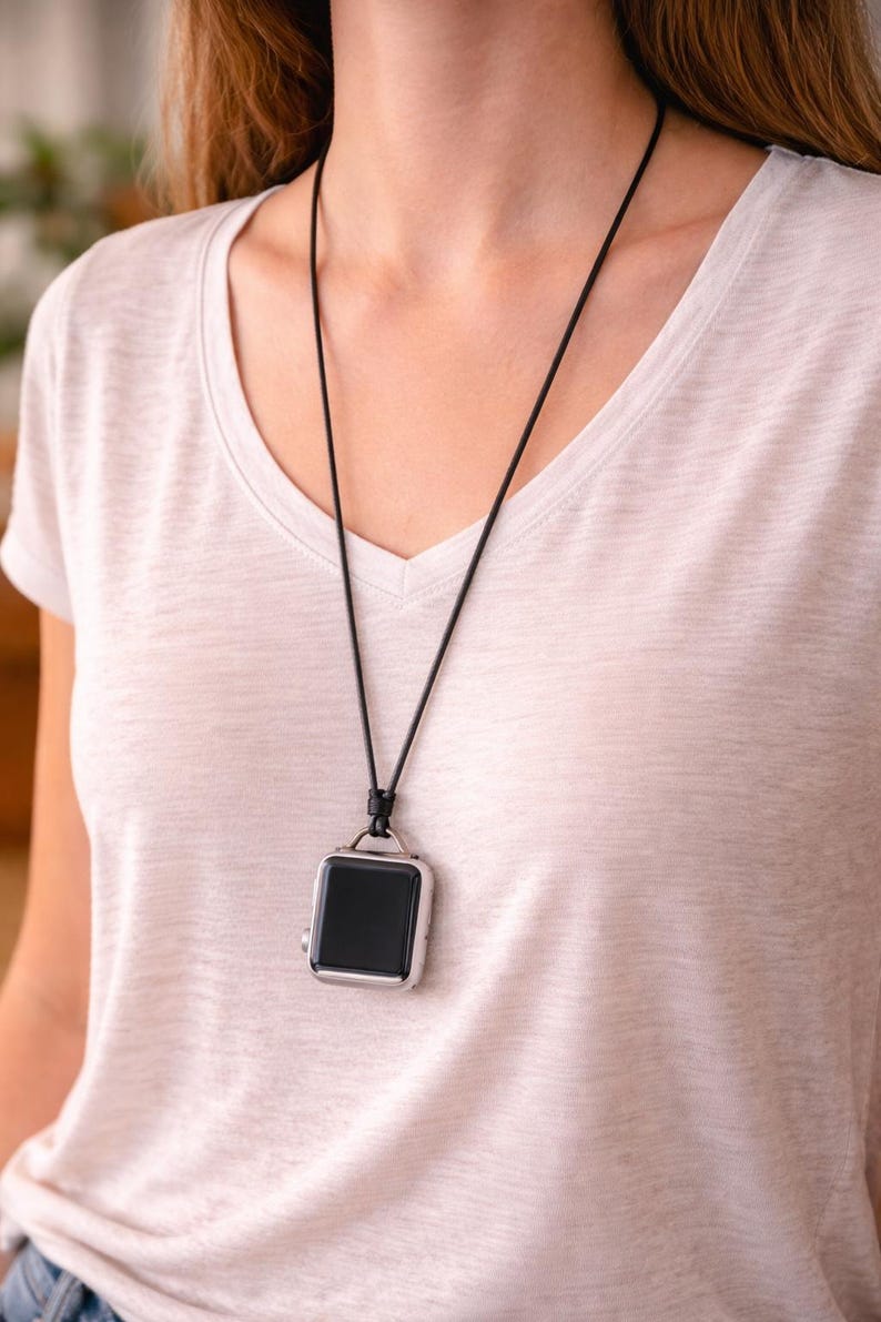 May include: A black smartwatch with a silver case hangs from a black cord necklace. The watch has a square face and is worn around the neck. The person is wearing a white v-neck top.