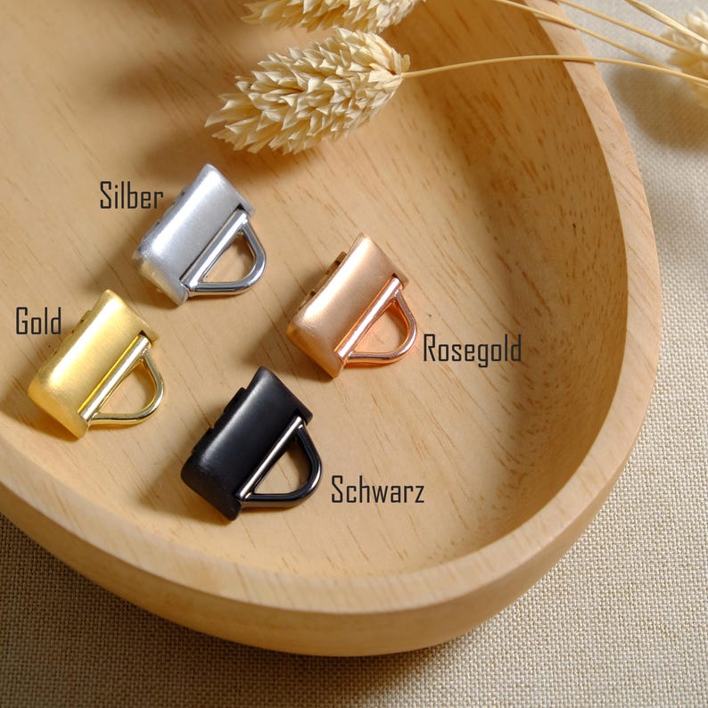 May include: Four metal purse hardware pieces in silver, gold, rose gold, and black are displayed. Each piece has a rectangular body with a D-ring. The word "Silber" is above the silver piece, "Gold" above the gold piece, "Rosegold" above the rose gold piece, and "Schwarz" above the black piece.