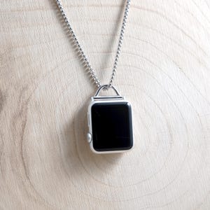 Könnte beinhalten: Eine silberne Apple Watch-Anhänger-Halskette. Das Zifferblatt der Uhr ist schwarz, das Gehäuse silberfarben. Die Halskette hat eine feine silberne Kugelkette. Die Uhr hängt an einer silbernen Schlaufe.