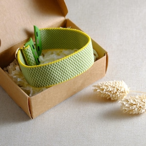 May include: A light green watch strap with a yellow trim, displayed in a brown cardboard box with white paper filler. The strap has a woven pattern. The watch strap is a light green colour.