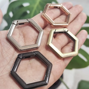 Op de afbeelding: Vier zeshoekige metalen karabijnhaakclips in zilver, goud, messing en zwart. De clips zijn open en klaar om aan een tas of ander item te bevestigen. De clips worden in een hand getoond.