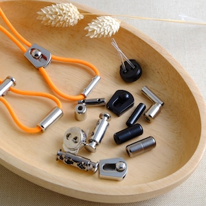 May include: Assortment of silver and black metal hardware for crafting, including cord ends, toggles, and stoppers. The hardware is displayed on a wooden plate.