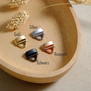 May include: A selection of metal strap buckles in gold, silver, rose gold, and black, arranged on a wooden tray. The buckles have a rectangular frame with a D-ring. The words "Gold", "Silber", "Rosegold", and "Schwarz" are printed on the image.