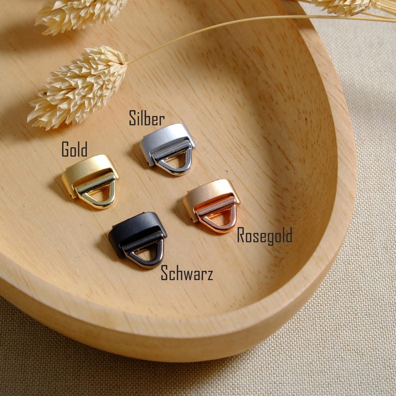 May include: A selection of metal strap buckles in gold, silver, rose gold, and black, arranged on a wooden tray. The buckles have a rectangular frame with a D-ring. The words "Gold", "Silber", "Rosegold", and "Schwarz" are printed on the image.
