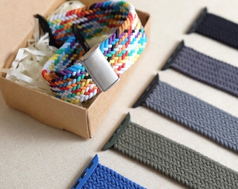 May include: A collection of braided elastic watch bands featuring one multicoloured woven strap inside a cardboard box and four additional solid-coloured straps in black, grey, olive green, and blue arranged in a row beside the box.