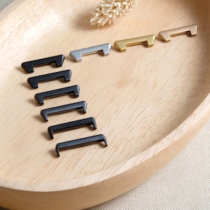 Könnte beinhalten: Eine Auswahl an Schrankgriffen in verschiedenen Metallausführungen, darunter Schwarz, Silber, Gold und Roségold, auf einem hellen Holztablett angeordnet. Die Griffe haben ein schlichtes, modernes Design mit einem gebogenen Griff und einer rechteckigen Basis. Ein getrockneter Weizenhalm ist im Hintergrund.