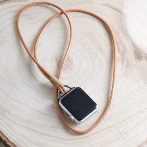 Peut inclure: Une montre connectée argentée avec un écran noir est attachée à un collier en cordon de cuir beige. Le cordon est bouclé et noué, conçu pour être porté autour du cou. La montre est présentée sur une surface en bois clair.