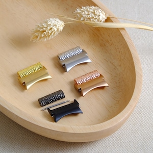 Puede incluir: Cuatro conectores de correa de reloj en diferentes acabados: oro, plata, oro rosa y negro. Cada conector tiene una superficie texturizada y una forma curva.
