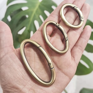 May include: Three antique brass oval snap hooks are displayed. The snap hooks are held in a hand, and are in various stages of being open or closed. The background is a green leaf.