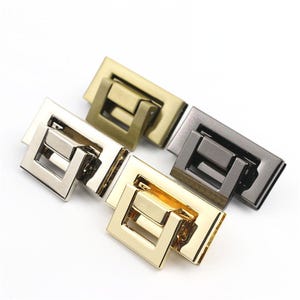 Op de afbeelding: Close-up van verschillende metalen portemonneesluitingen in verschillende afwerkingen: goud, zilver, brons en gunmetal. Elke sluiting heeft een rechthoekige basis met een vierkante, scharnierende sluiting. De sluitingen zijn gerangschikt op een wit oppervlak.