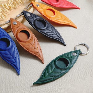 May include: A collection of leaf-shaped keychains in various colors, including blue, brown, black, orange, red, and green. Each keychain features a circular opening and a silver keyring. The keychains have a quilted leaf pattern.