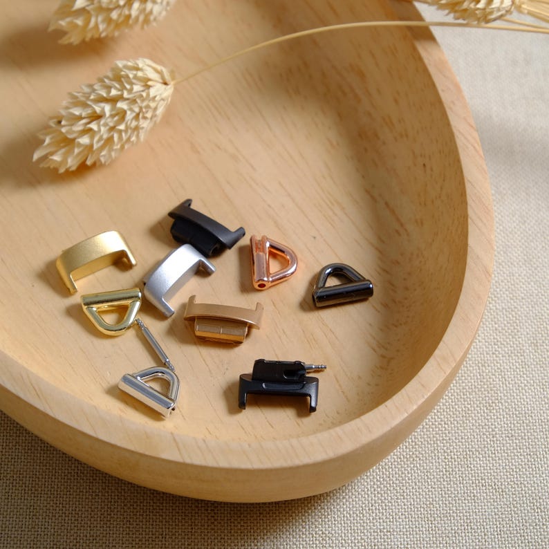 May include: Assortment of metal watch band connectors in various colors, including black, silver, gold, and rose gold. The connectors are displayed on a wooden surface.