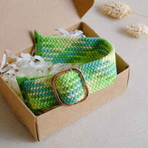 Könnte beinhalten: Ein geflochtenes Armband in Grün, Blau und Weiß mit einer goldenen Metallschnalle. Das Armband befindet sich in einer Schachtel mit weißem Seidenpapier.