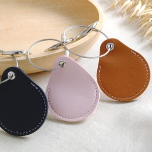 May include: Three leather keychains in navy, pink, and brown, each with a silver-tone metal wire loop and clasp. The teardrop-shaped tags have white stitching around the edges. The keychains are displayed on a light-colored surface.