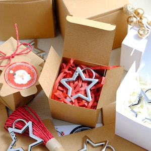 Puede incluir: Tres pequeñas cajas de regalo con papel marrón y blanco. Las cajas están llenas de papel de seda rojo y blanco. Cada caja contiene un llavero en forma de estrella plateada.