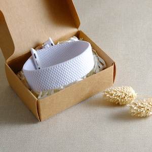 Könnte beinhalten: Ein weißes, strukturiertes Uhrenarmband liegt in einer offenen, braunen Pappschachtel, die mit weißem Papier gefüllt ist. Das Armband hat ein sich wiederholendes Muster aus kleinen, erhabenen Punkten. Getrocknete Blumen sind im Hintergrund.
