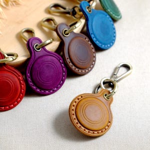 May include: A collection of colorful leather keychains in red, purple, brown, blue, and green. Each keychain features a circular design with a stitched border and a metal clasp for attaching to keys or bags. The keychains are made of leather.