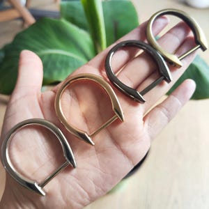 Op de afbeelding: Vier metalen D-ringen in zilver, goud, gunmetal en messing tinten. Elke ring heeft een afgeronde bovenkant en een rechte staaf aan de onderkant, met een puntig uiteinde. De ringen worden in een hand gehouden, met een groene plant op de achtergrond.