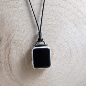 May include: A silver smartwatch with a black screen hangs from a black cord necklace. The watch has a square face with rounded corners and a silver metal loop. The background is a light brown wooden surface.