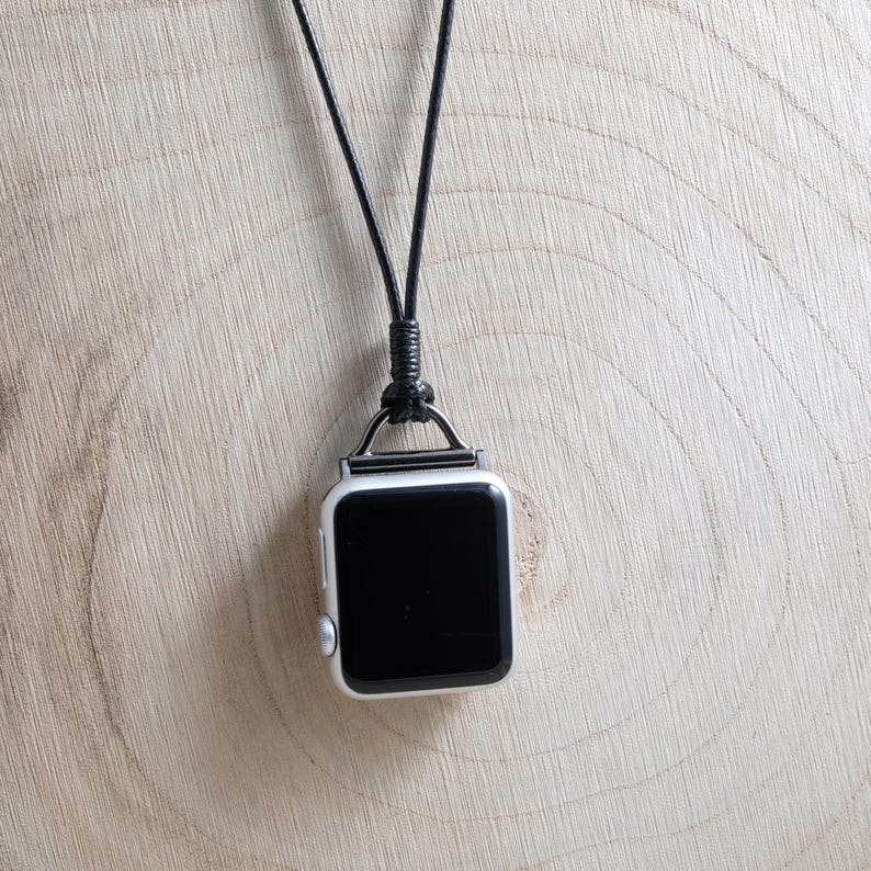May include: A silver smartwatch with a black screen hangs from a black cord necklace. The watch has a square face with rounded corners and a silver metal loop. The background is a light brown wooden surface.