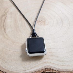 May include: A silver smartwatch with a black screen hangs from a black cord, forming a necklace. The watch is square and has a silver bezel. The cord is knotted at the top, and the background is a light-coloured wooden surface.