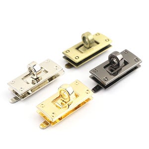 May include: Four metal purse clasps in gold, silver, and gunmetal finishes. Each clasp has a rectangular base with rounded corners and a circular loop. The clasps are designed for bags and other craft projects.