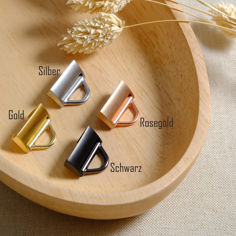 May include: Four metal purse hardware pieces in gold, silver, rose gold, and black, each with a rectangular bar and a D-ring. The pieces are displayed in a wooden bowl with dried wheat stalks.