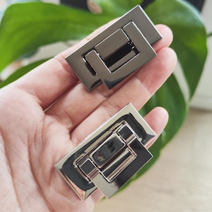 Op de afbeelding: Twee rechthoekige, gunmetal-kleurige portemonneesluitingen. Elke sluiting heeft een rechthoekig frame met een scharnierende, kleinere rechthoek in het midden. De sluitingen zijn glanzend en reflecteren het licht. De achtergrond is een wazig groen blad.