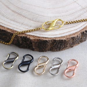 Pode incluir: Um colar de corrente tipo caixa em tom dourado com um fecho lagosta em forma de S. Abaixo do colar, cinco fechos adicionais em forma de S estão dispostos em fila, apresentando acabamentos em latão antigo, preto fosco, ouro, prata e ouro rosa.