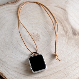May include: A silver smartwatch with a black screen is suspended from a tan leather cord, knotted at the end. The watch is displayed on a light-colored wooden surface, showcasing a minimalist design.