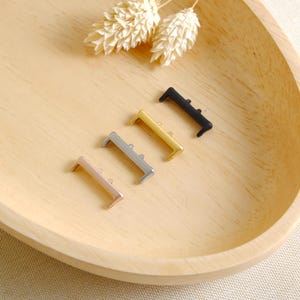 Puede incluir: Cuatro cierres de barra metálicos en oro rosa, plata, dorado y negro, dispuestos sobre una bandeja de madera. Cada cierre tiene forma rectangular con un extremo curvado. Los cierres son pequeños, probablemente para joyería o manualidades.