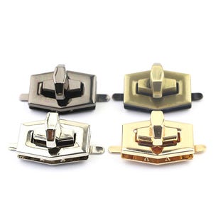 Könnte beinhalten: Vier Metall-Geldbörsenverschlüsse in verschiedenen Ausführungen: Gunmetal, Antik-Messing, Silber und Gold. Jeder Verschluss hat einen rechteckigen Körper mit einem zentralen Hebel und kleinen seitlichen Laschen zur Befestigung. Geeignet für Taschen.