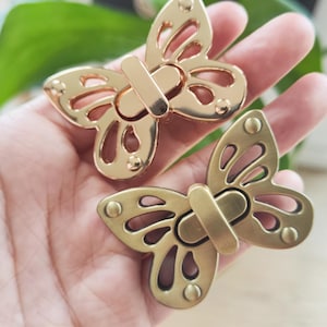 Op de afbeelding: Twee goudkleurige vlindervormige sluitingen. De sluitingen hebben een decoratief vlinderontwerp met uitgesneden details. Elke sluiting heeft een rechthoekige staafsluiting. De sluitingen zijn van metaal, waarschijnlijk voor een handtas of knutselproject.