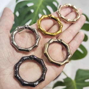 Pode incluir: Uma coleção de cinco anéis de metal com design de bambu em vários acabamentos: prata, ouro, bronze e preto. Os anéis são abertos e projetados para serem usados como fechos ou conectores. O fundo inclui folhas verdes.