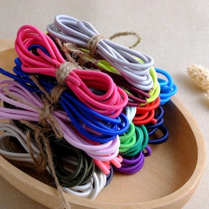 Könnte beinhalten: Eine Sammlung bunter elastischer Kordeln, gebündelt und mit Bindfaden zusammengebunden, in einer Holzschale angeordnet. Die Kordeln sind in verschiedenen Farben erhältlich, darunter Rosa, Blau, Grün und Weiß. Die Kordeln sind eng gewickelt und scheinen neu zu sein.