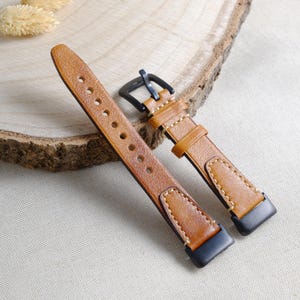 Puede incluir: Dos correas de reloj de cuero marrón con hebillas negras. Las correas presentan costuras visibles y varios orificios de ajuste. Las correas se muestran sobre una superficie clara.