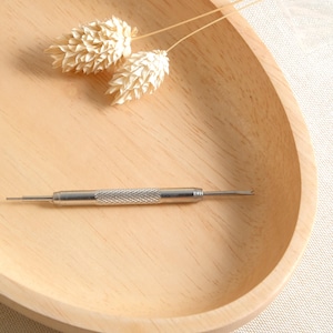 Puede incluir: Una herramienta de reparación de correa de reloj plateada sobre una superficie de madera. La herramienta tiene un extremo puntiagudo y un extremo plano.