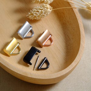 Puede incluir: Cuatro cierres de anillo en D de metal en plata, oro, oro rosa y negro. Los cierres se muestran sobre una superficie de madera.