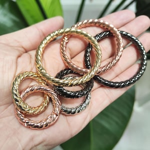 Puede incluir: Una colección de anillos metálicos en varios colores, incluyendo dorado, oro rosa, plateado y negro. Cada anillo presenta un diseño de cuerda trenzada. Los anillos se sostienen en una mano, mostrando sus diferentes acabados metálicos.