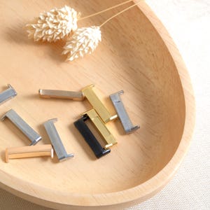 Puede incluir: Surtido de cierres de correa de reloj de metal en dorado, plateado y negro. Los cierres est&aacute;n dispuestos sobre una superficie de madera.