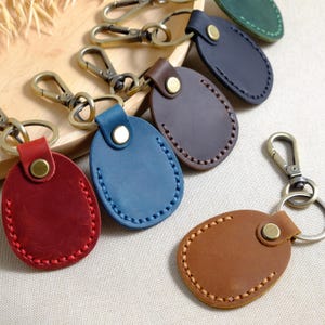 May include: A collection of leather keychains in various colors, including red, blue, brown, and navy. Each keychain has a stitched oval design and a metal clasp. The keychains are displayed on a wooden surface.