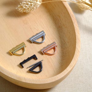 Könnte beinhalten: Eine Sammlung von Metallbeschlägen für Handtaschen in Gold, Silber, Roségold und Schwarz. Die Beschläge umfassen rechteckige Stangen mit D-Ring-Design. Die Artikel werden in einer Holzschale auf einer neutralen Oberfläche präsentiert.