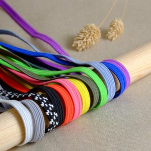 Könnte beinhalten: Eine Sammlung bunter Gummibänder in verschiedenen Farben, darunter Lila, Blau, Grün, Gelb, Rosa, Orange, Schwarz und Weiß. Die Gummibänder sind fächerförmig auf einer Holzoberfläche angeordnet.