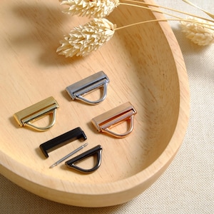 May include: Four metal D-ring hardware pieces in different finishes: gold, silver, black, and rose gold. The pieces are arranged on a wooden surface.