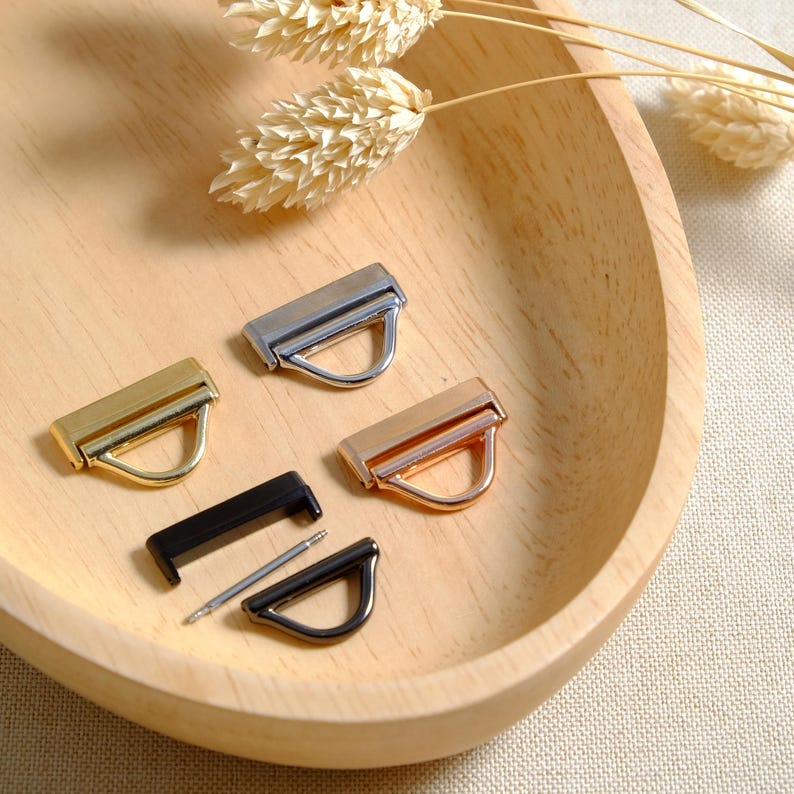 May include: Four metal D-ring hardware pieces in different finishes: gold, silver, black, and rose gold. The pieces are arranged on a wooden surface.