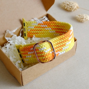 Puede incluir: Una banda trenzada para Apple Watch de color amarillo y blanco con una hebilla de metal dorado. La banda está en una caja de cartón marrón con papel de seda blanco.